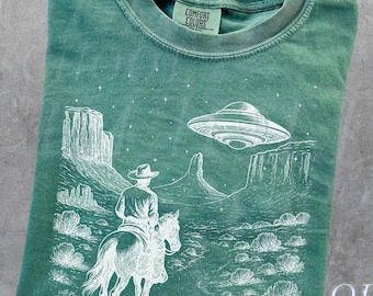 May include: Light green t-shirt with a white graphic of a cowboy on a horse, a UFO, and desert landscape. The text "OLD SCHOOL CULT" and "Light Green" are also visible.