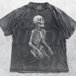 May include: Pepper-colored t-shirt featuring a detailed white skeleton graphic. The design is centered on the front of the shirt, with the skeleton's rib cage and limbs visible. The shirt has a vintage, worn look.