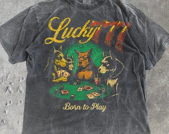 May include: Dark gray t-shirt with a graphic of dogs playing poker. The text "Lucky 77" is above the dogs, and "Born to Play" is below. The shirt has a vintage, distressed look.