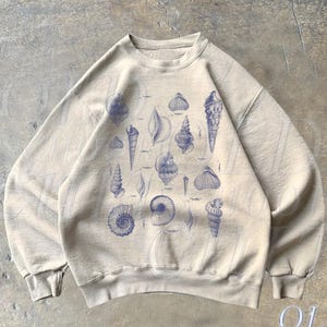 May include: A beige sweatshirt with a blue print of various seashells. The print is centered on the front of the sweatshirt.