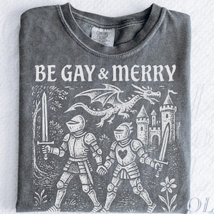 Puede incluir: Camiseta color pimienta con el texto "BE GAY & MERRY" sobre un gr&aacute;fico blanco de dos caballeros, un drag&oacute;n y un castillo. El dise&ntilde;o tiene un aspecto vintage y desgastado. La camiseta tiene cuello redondo y mangas cortas.