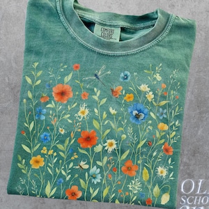 May include: Light green t-shirt with a floral design featuring orange, blue, and yellow flowers, green stems, and a dragonfly. The shirt has a crew neck and the words "OLD SCHOOL CULT" are printed on the bottom right.