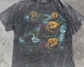 May include: A dark gray t-shirt featuring a graphic of otters in water, with sea urchins and aquatic plants. The shirt has a vintage, worn look. The text "OLD SCHOOL CULT" is printed on the lower right.
