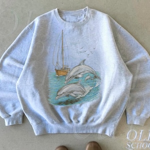 Puede incluir: Sudadera gris claro con un gráfico de dos delfines saltando de las olas del océano, con un velero en el fondo. El diseño incluye pájaros en el cielo. La sudadera tiene cuello redondo y mangas largas. El texto "OLD SCHOOL CULT" está impreso en la parte inferior derecha.
