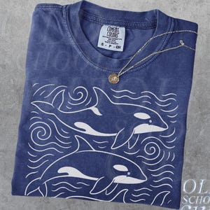 Puede incluir: Una camiseta azul marino con un gráfico blanco de dos orcas nadando en el océano. La camiseta está doblada y tiene una etiqueta Comfort Colors. Un collar dorado con un colgante descansa sobre la camiseta. El texto "True Navy" y "OLD SCHOOL CULT" son visibles.