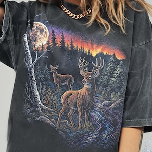 Könnte beinhalten: Dunkelgraues T-Shirt mit einer Waldlandschaft, die zwei Hirsche, einen Vollmond und einen farbenfrohen Sonnenuntergang zeigt. Das Shirt hat kurze Ärmel und eine lockere Passform. Das Wort "Pepper" ist unten links aufgedruckt.