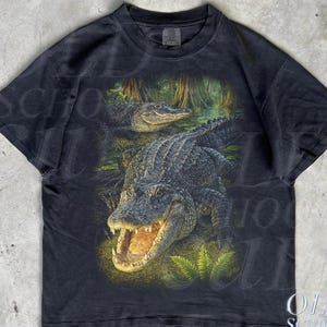 May include: Black t-shirt with a detailed graphic of two alligators in a swamp. The larger alligator is in the foreground with its mouth open. The shirt has a vintage appearance.