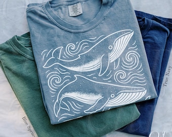 May include: Three t-shirts in blue, green, and navy. The blue shirt features a white graphic of two whales and swirling water. The green shirt is labeled "Blue Spruce" and the navy shirt is labeled "True Navy". The bottom left shirt is labeled "Blue Jean".