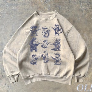 May include: A tan sweatshirt with a vintage-style print of various animals playing musical instruments. The animals include a bear, raccoon, and a squirrel. The text "OLD SCHOOL CULT" is printed on the bottom right corner of the sweatshirt.