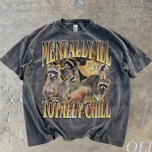 以下が含まれることがあります： グレーのTシャツに、アライグマとカピバラのグラフィックプリントが施されています。Tシャツには「Mentally Ill Totally Chill」というテキストが黄色でプリントされています。