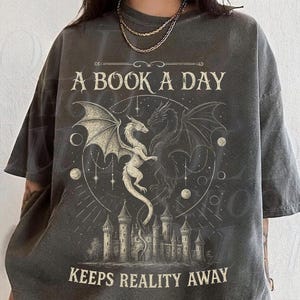 May include: Charcoal grey t-shirt with a fantasy graphic. The design includes two dragons, a castle, and the text "A BOOK A DAY KEEPS REALITY AWAY". The shirt has a vintage, worn appearance.