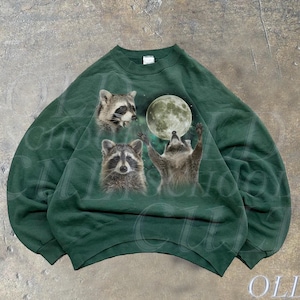 Op de afbeelding: Een donkergroen sweatshirt met een grafische print van drie wasberen die naar een volle maan kijken. De maan is lichtgroen en de wasberen zijn bruin en grijs. Het sweatshirt heeft een ronde hals en lange mouwen.