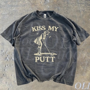 May include: A black t-shirt with a vintage-style graphic of a woman putting a golf ball. The graphic is in a cream color and says "Kiss My Putt".