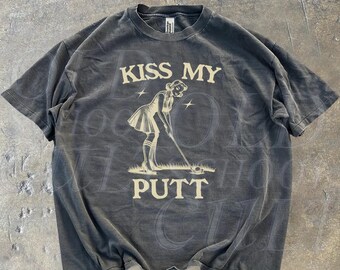 May include: A black t-shirt featuring a vintage-style graphic of a woman putting a golf ball. The graphic is in a cream colour and says "Kiss My Putt".