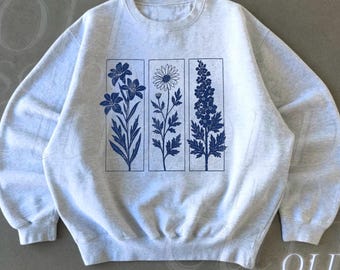 May include: A light gray sweatshirt with a blue floral design. The design features three vertical rectangular panels, each with a different botanical illustration. The words "OLD SCHOOL CULT" are printed in white on the lower right.