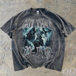 K&ouml;nnte beinhalten: Ein graues T-Shirt mit einem Grafikdruck von zwei W&ouml;lfen, die zum Mond heulen. Der Text "GOT THAT DOG IN ME" ist in gotischer Schrift unter den W&ouml;lfen gedruckt.