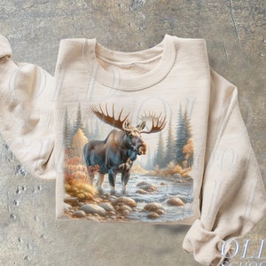 May include: A beige sweatshirt with a graphic print of a moose standing in a stream. The moose is brown with large antlers. The background is a forest scene with trees and rocks.