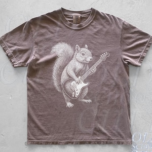 Könnte beinhalten: Espressofarbenes T-Shirt mit einer detaillierten Illustration eines Eichhörnchens, das E-Gitarre spielt. Das Eichhörnchen ist in Grau- und Weißtönen dargestellt, mit einem flauschigen Schwanz und konzentriertem Ausdruck. Das Shirt hat einen klassischen Rundhalsausschnitt und kurze Ärmel.
