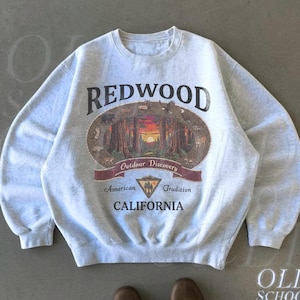 May include: Ash-colored sweatshirt with the word "REDWOOD" in large, dark letters above an oval graphic of a forest scene. The graphic includes the text "Outdoor Discovery", "American Tradition", and "CALIFORNIA".
