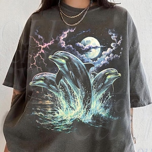 Könnte beinhalten: Dunkelgraues T-Shirt mit einer Grafik von drei Delfinen, die aus dem Wasser springen, unter einem Vollmond, mit Blitzen und Wolken im Hintergrund. Das T-Shirt hat einen Vintage-Look.