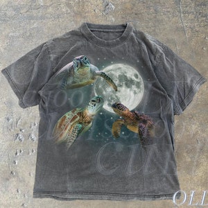 May include: A dark grey t-shirt featuring a graphic print of three sea turtles swimming around a full moon. The text "OLD SCHOOL CULT" is printed in white at the bottom of the shirt.