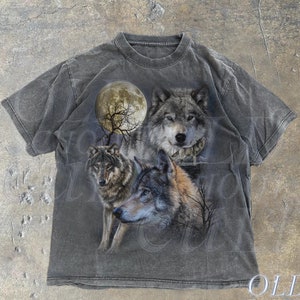 May include: A gray t-shirt with a vintage look featuring a graphic of three wolves howling at the moon. The wolves are depicted in a realistic style with detailed fur and expressions. The moon is a full moon with a light gray color.