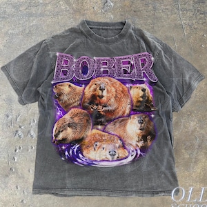 Könnte beinhalten: Ein graues T-Shirt mit einem verblassten Look, das eine violette Grafik mit dem Wort "BOBER" zeigt, umgeben von Cartoon-Bibern.
