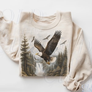Peut inclure: Sweat-shirt couleur sable avec un motif détaillé d'un aigle à tête blanche en vol au-dessus d'une forêt et d'une cascade. L'aigle a des plumes marron et blanches, avec un bec jaune. Le motif comprend des arbres et des montagnes.
