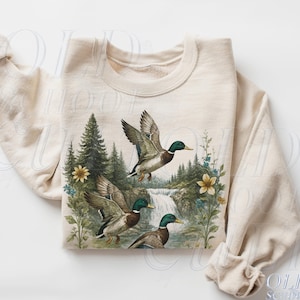 Peut inclure: Sweat-shirt couleur sable avec une scène de nature représentant trois canards volant au-dessus d'une cascade et d'une forêt. Les canards ont la tête verte et le corps brun. Des fleurs jaunes et bleues parsèment le motif.