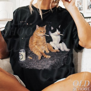 May include: Black t-shirt with a graphic of two cats smoking cigarettes, a can, and cigarette butts. The shirt has the text "OLD SCHOOL CULT" and "Black" printed on it. The model is 5'8" and wearing XL.