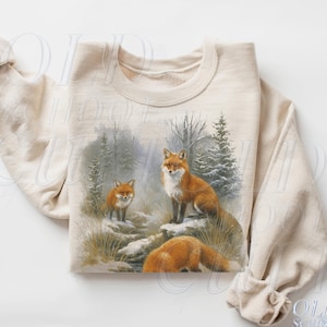 May include: Sand-colored sweatshirt featuring a detailed illustration of three foxes in a winter forest scene. The foxes have reddish-orange fur and are set against a backdrop of snow-covered ground and evergreen trees. The text "OLD SCHOOL CULT" is visible.