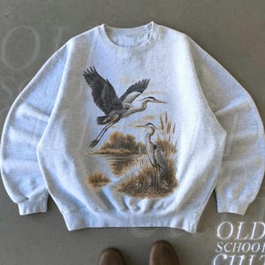 May include: Ash-colored sweatshirt featuring a nature scene with two herons, one in flight and the other standing near a body of water. The sweatshirt has a crew neck and long sleeves. The text "OLD SCHOOL CULT" is printed on the bottom right.