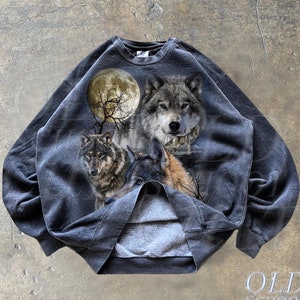 Peut inclure: Sweat-shirt gris foncé avec un motif détaillé de loups sous une pleine lune. Le design comprend trois loups avec des textures de fourrure réalistes et un fond de lune et d'arbres dénudés. Le sweat-shirt a un aspect vintage et usé, avec le texte "OLD SCHOOL CULT" en bas.