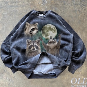 Op de afbeelding: Een donkergrijs sweatshirt met een grafische print van drie wasberen die naar een volle maan reiken. Het sweatshirt heeft een ronde hals en lange mouwen.
