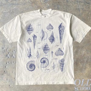 May include: A white t-shirt with a blue print of various seashells. The print includes a variety of shapes and sizes of seashells.