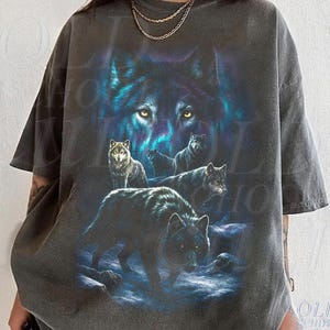 以下が含まれることがあります： 詳細なオオカミのグラフィックが特徴の濃い灰色のTシャツ。デザインには、輝く黄色の目を持つ大きなオオカミの顔と、前景にいくつかのオオカミが含まれています。このシャツはヴィンテージで、使い古された外観をしています。