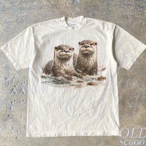Op de afbeelding: Een wit T-shirt met een grafische print van twee otters in een rivier. De otters zijn bruin en hebben een speelse uitdrukking. Het shirt heeft een ronde hals en korte mouwen.