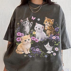 Può includere: T-shirt grigio scuro con un design floreale con cinque gattini di vari colori. I gattini sono circondati da fiori viola e rosa, con una farfalla rosa e il testo "OLD SCHOOL CULT".