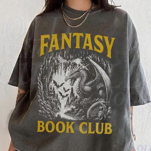 Pode incluir: T-shirt cinza escuro com as palavras "FANTASY BOOK CLUB" em amarelo. A camisa apresenta um gráfico detalhado de um dragão em uma caverna com morcegos. A camisa tem um ajuste folgado.