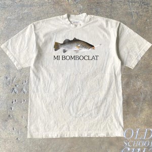 Könnte beinhalten: Ein weißes T-Shirt mit einem Aufdruck eines Fisches, der eine Zigarette raucht. Unter dem Fisch steht der Text "MI BOMBOCLAT".