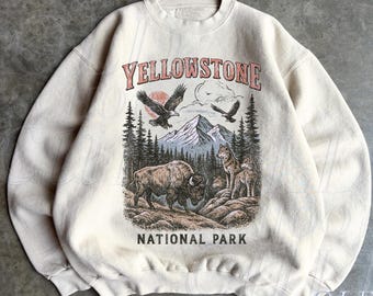 May include: Sand-colored sweatshirt featuring a graphic of Yellowstone National Park. The design includes a bison, wolves, eagles, mountains, and the text "YELLOWSTONE NATIONAL PARK." The sweatshirt has a crew neck and long sleeves.