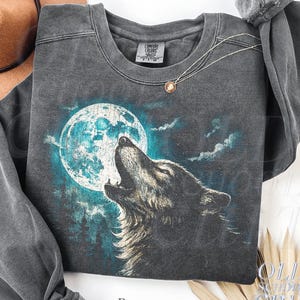 May include: A dark gray sweatshirt featuring a wolf howling at a full moon. The graphic is in shades of blue, white, and gray. The sweatshirt has a crew neck and long sleeves. The text "OLD SCHOOL CULT" is visible on the bottom right.