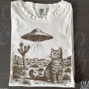 May include: Ivory t-shirt with a desert scene graphic. The design features a cat, a UFO, and a desert landscape with a Joshua tree and mountains. The t-shirt has a crew neck and the text "OLD SCHOOL CULT".