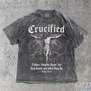 May include: Charcoal grey t-shirt with a graphic of a crucified figure and the word "Crucified". The text "Father, forgive them: for they know not what they do. - Luke 23:34" is also printed on the shirt. The t-shirt has a vintage, worn appearance.