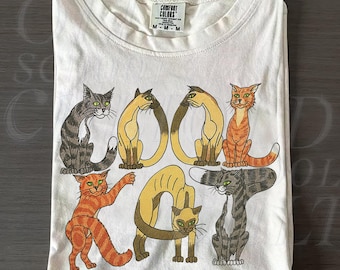 Op de afbeelding: Ivoor kleurig t-shirt met een afbeelding van gestileerde katten die de woorden "Cool Kat" vormen. De katten hebben verschillende poses en kleuren, waaronder oranje, grijs en bruin. Het shirt is netjes opgevouwen.