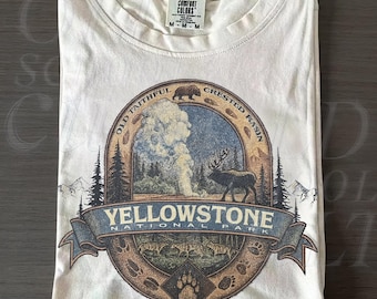 May include: Ivory-colored t-shirt featuring a circular graphic of Yellowstone National Park. The design includes a geyser, elk, bear, and the text "Yellowstone National Park." The shirt is folded and has a Comfort Colors tag.