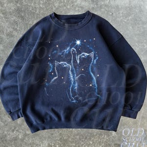 May include: Navy blue sweatshirt featuring a celestial design with two cats gazing at a star. The cats are depicted in a soft, blue hue against a starry background. The sweatshirt has a classic crew neck and long sleeves.
