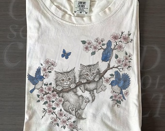 May include: Ivory-colored t-shirt featuring a graphic of two kittens on a flowering branch with blue birds and butterflies. The shirt has a crew neck and a tag that reads "COMFORT COLORS". The text "OLD SCHOOL CULT" is in the bottom right corner.