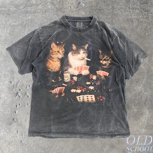 Op de afbeelding: Donkergrijs t-shirt met een pokerscène met drie katten. Eén kat rookt een sigaret terwijl hij kaarten vasthoudt. Het shirt heeft de tekst "Old School Cult" en "Pepper" erop gedrukt.