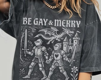 May include: Dark gray t-shirt with the text "BE GAY & MERRY" in white. The graphic features two knights, a dragon, and a castle. The shirt has short sleeves and a relaxed fit.